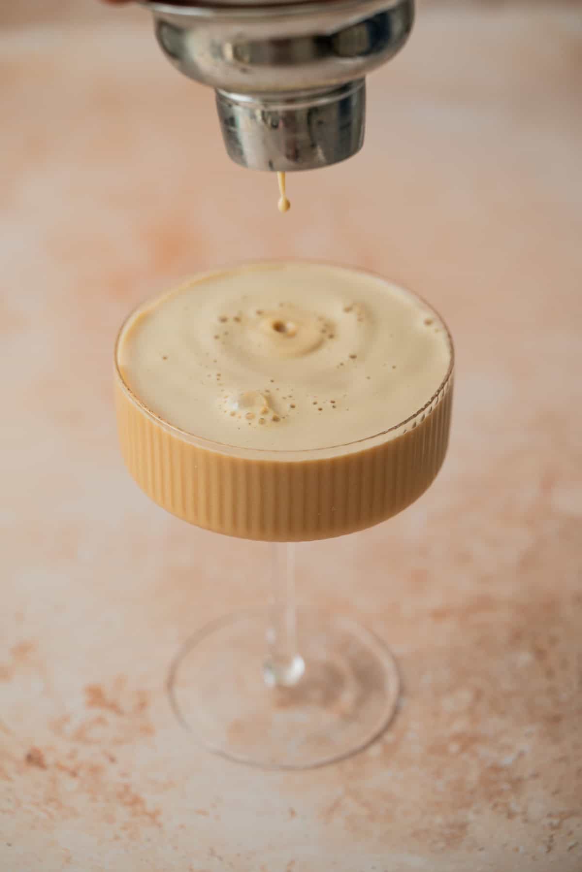 A creamy, frothy Eggnog Espresso Martini is poured into a coupe glass from a shaker, with a drop just about to fall. The drink has a light brown color and is set against a light, textured background.