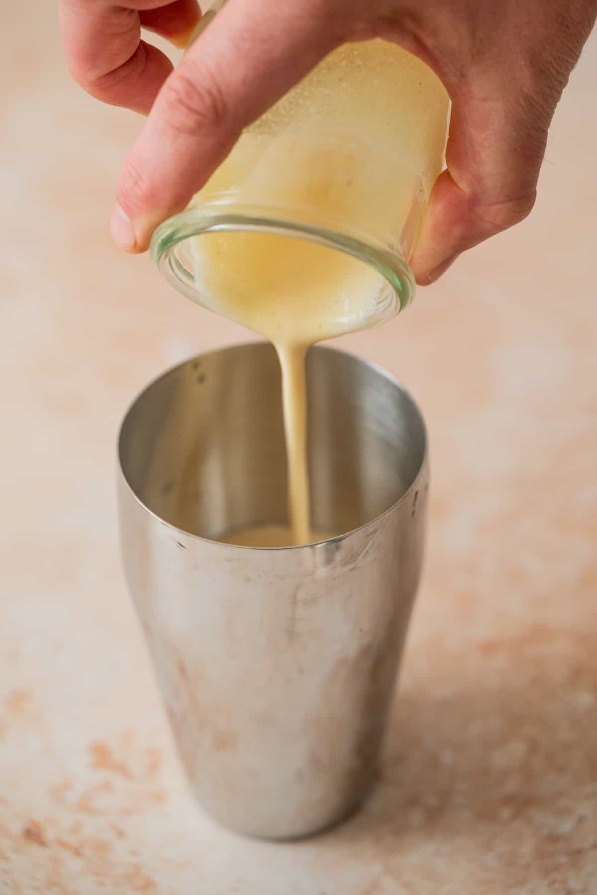 A hand pours a thick, yellow liquid from a glass jar into a metal cocktail shaker on a light, textured surface—perfect for crafting an Eggnog Espresso Martini.