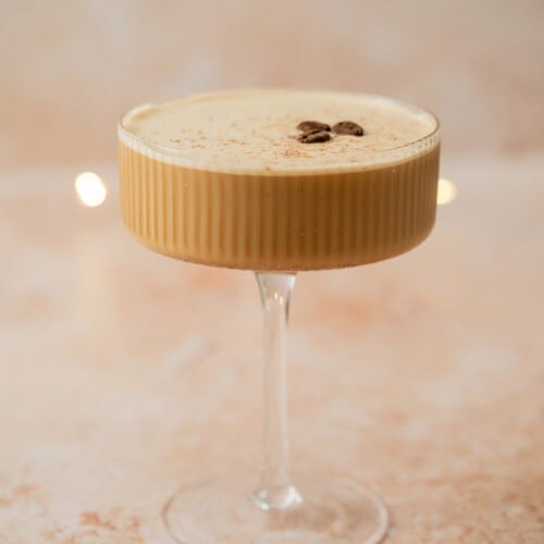 A creamy Eggnog Espresso Martini in a ribbed coupe glass, topped with two coffee beans and a dusting of cocoa powder, set on a light beige surface with a soft, blurred background.