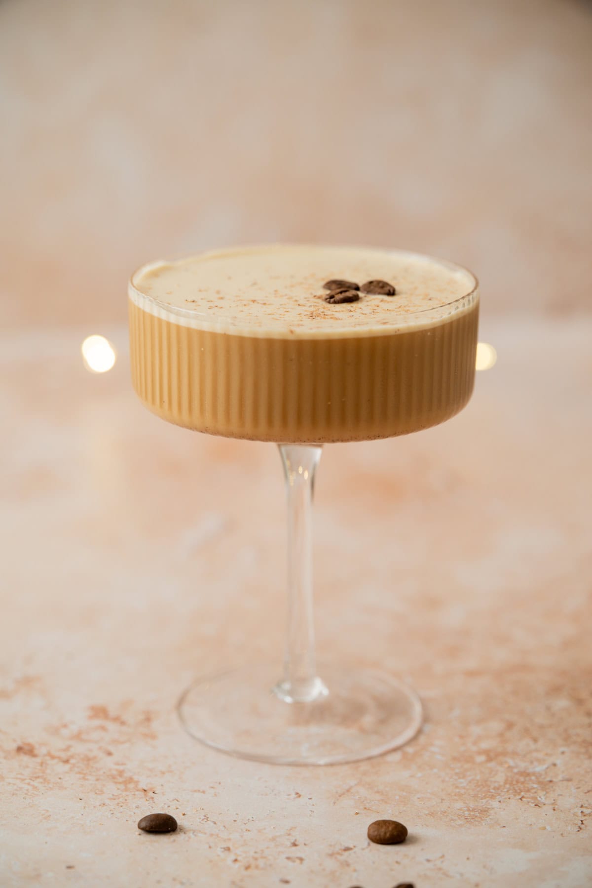 A creamy Eggnog Espresso Martini in a ribbed coupe glass, topped with two coffee beans and a dusting of cocoa powder, set on a light beige surface with a soft, blurred background.
