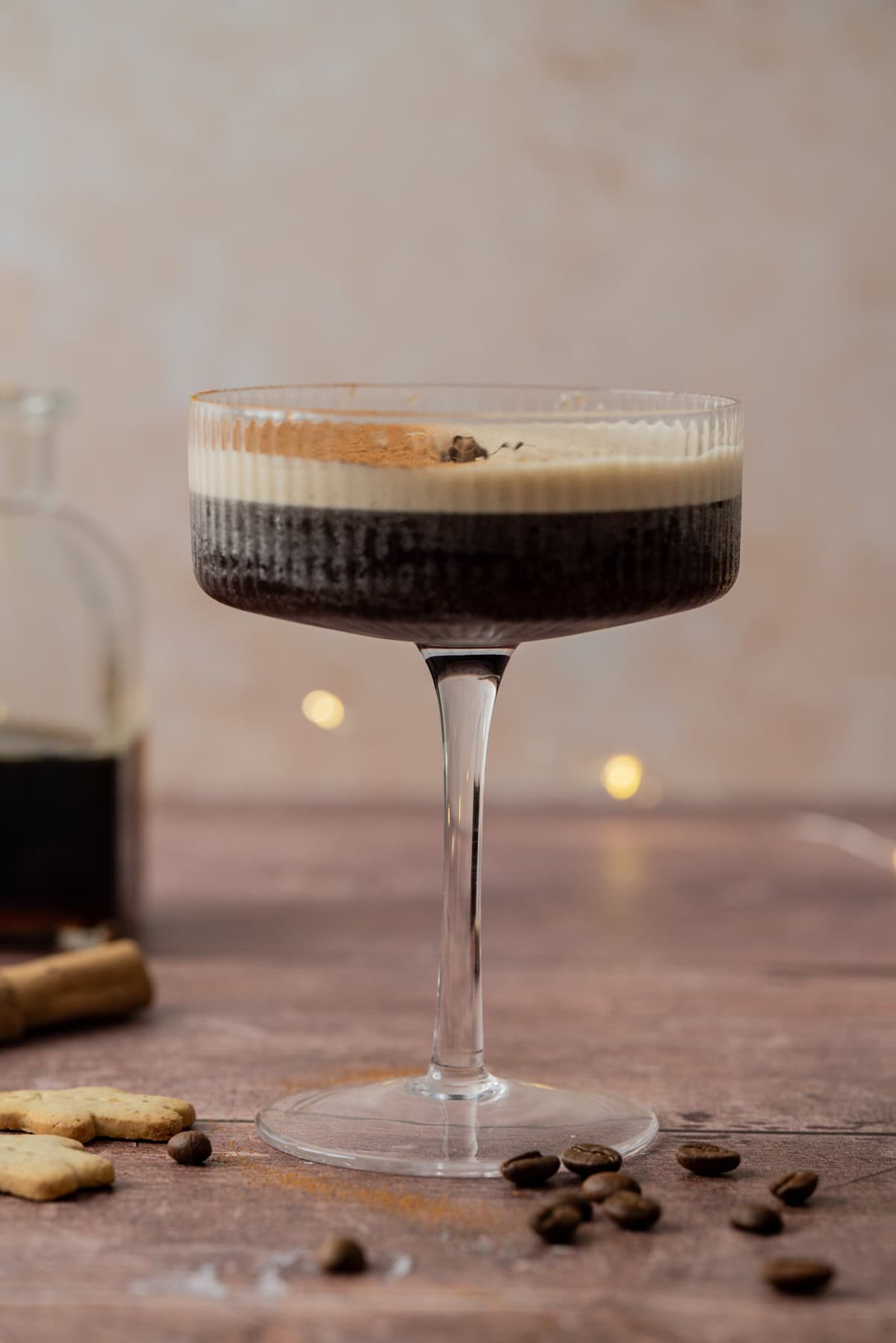 A glass of gingerbread espresso martini with a creamy foam layer on top, garnished with coffee beans, sits on a wooden surface dotted with scattered coffee beans. Blurred lights and a bottle appear in the background.