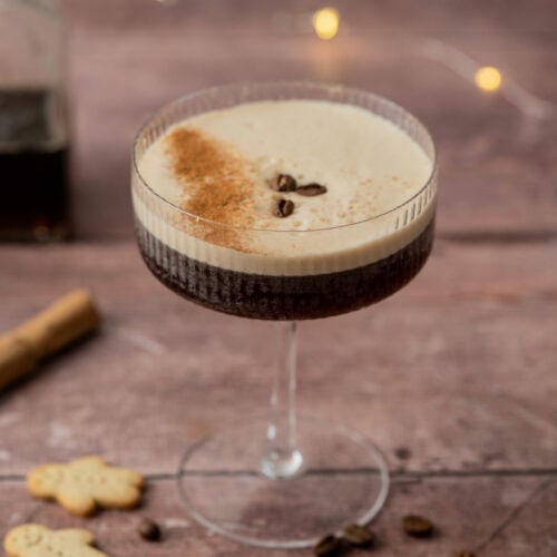 A coupe glass filled with a frothy gingerbread espresso martini, topped with three coffee beans and a dusting of cocoa. Coffee beans, gingerbread cookies, and a cinnamon stick are scattered on the rustic wooden surface.