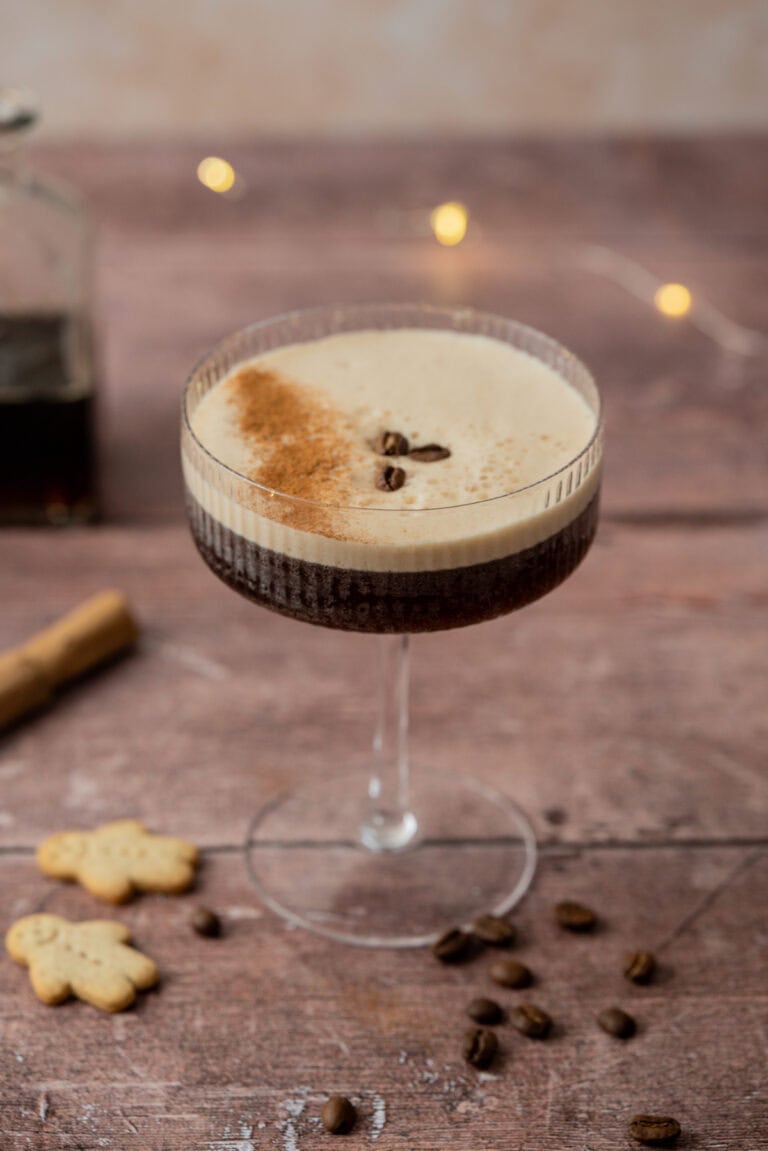 A coupe glass filled with a frothy gingerbread espresso martini, topped with three coffee beans and a dusting of cocoa. Coffee beans, gingerbread cookies, and a cinnamon stick are scattered on the rustic wooden surface.
