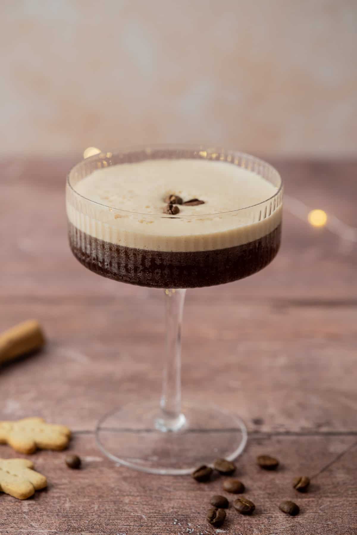 A martini glass filled with a frothy gingerbread espresso martini, garnished with three coffee beans, sits on a wooden surface with scattered coffee beans and cookies nearby.