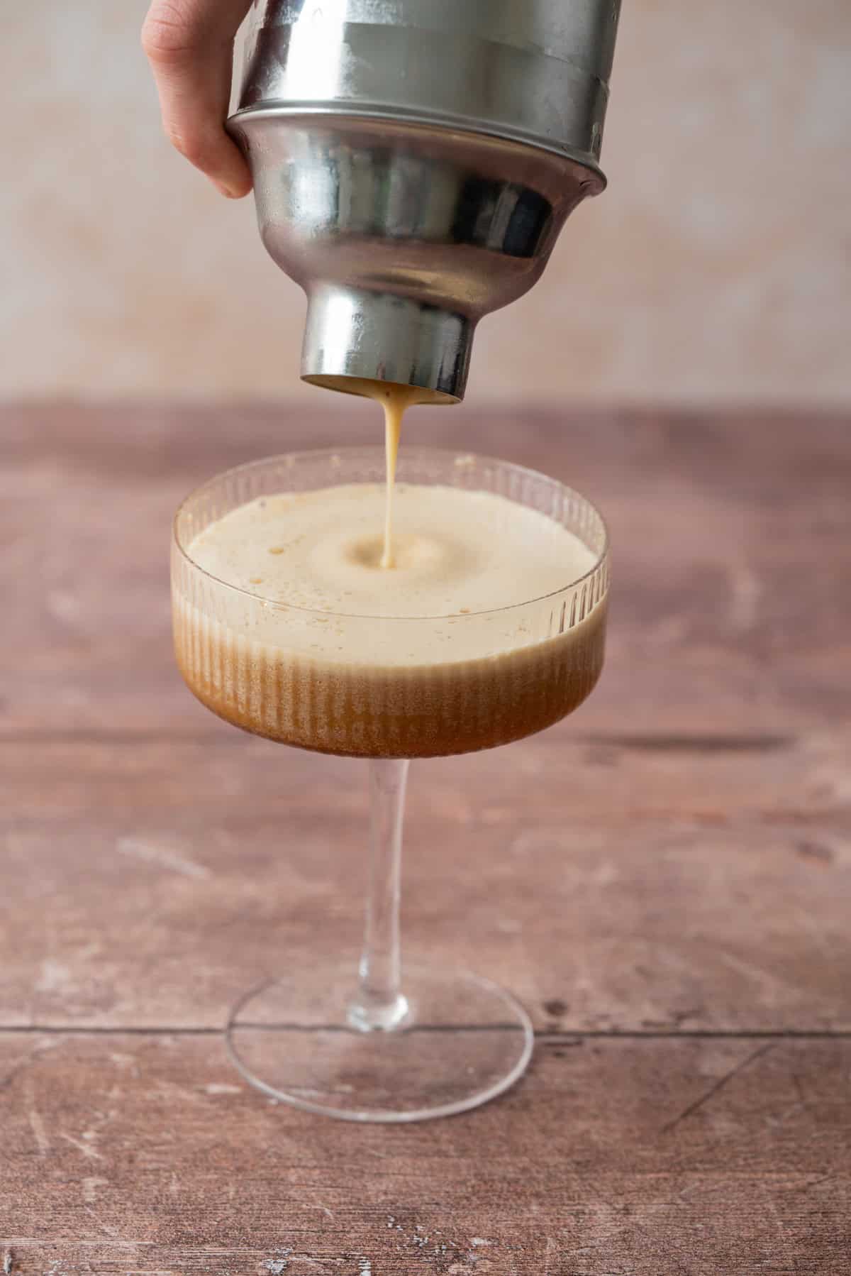 A hand pours a creamy, frothy gingerbread espresso martini from a cocktail shaker into a coupe glass on a wooden surface.