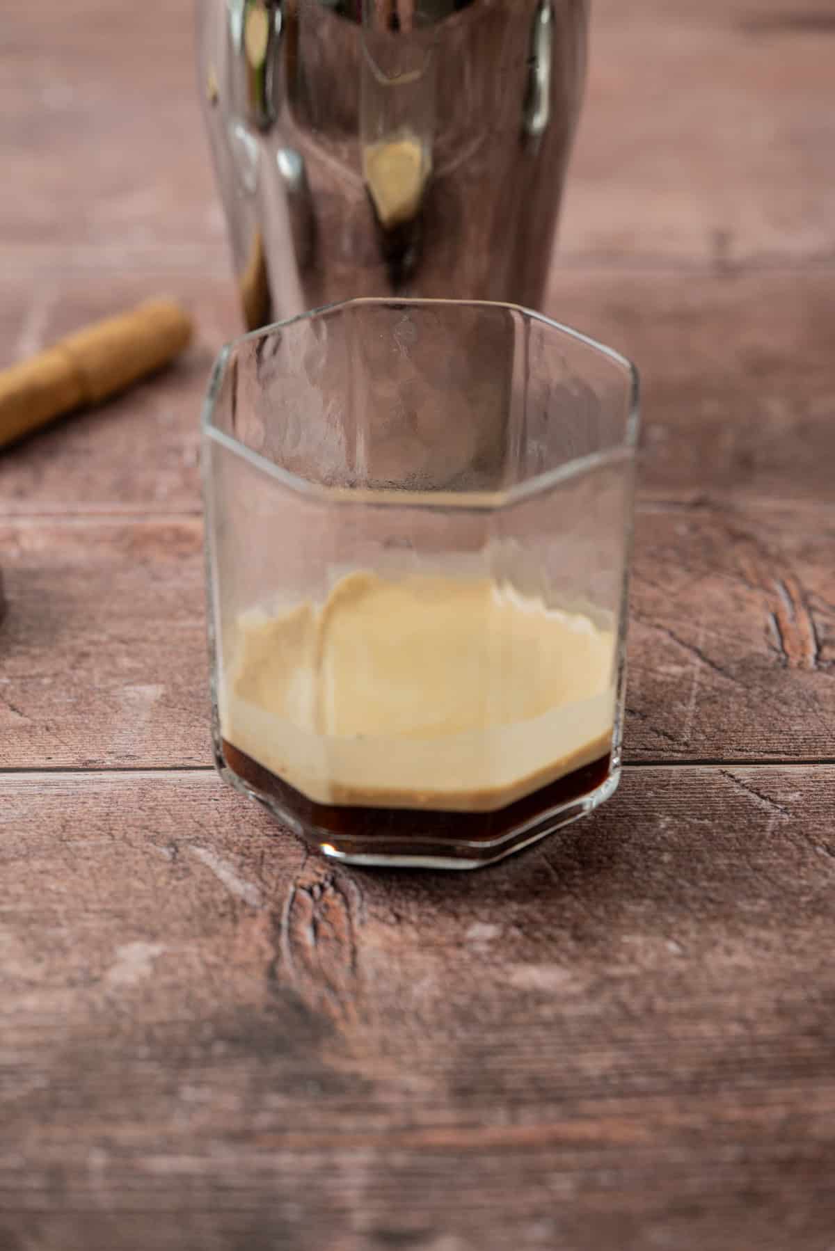 A hexagonal glass with a small amount of espresso and crema sits on a rustic wooden surface, with a blurred shaker and biscuit in the background.