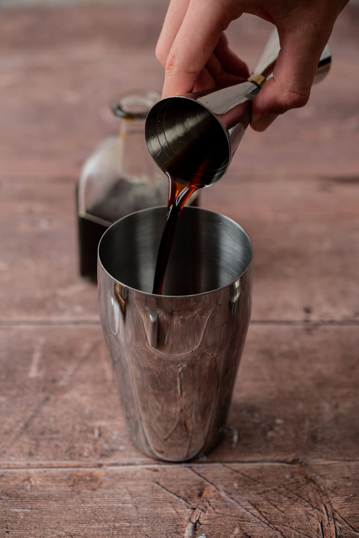 A hand pours a dark liquid from a jigger into a metal cocktail shaker, crafting a gingerbread espresso martini. A brown bottle rests in the background on a wooden surface.