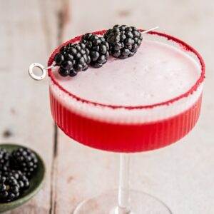 A vibrant red cocktail in a glass with a sugared rim, garnished with three blackberries on a skewer. The drink is frothy on top. A small green plate with extra blackberries is in the background on a wooden surface.