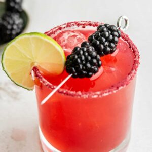 A vibrant red cocktail garnished with a slice of lime on the glass rim, and skewered blackberries on top. The rim is coated with a reddish sugar. The drink is served in a clear glass with ice cubes.
