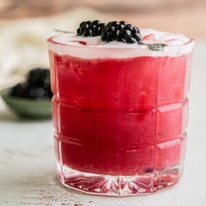 A refreshing blackberry whisky sour cocktail garnished with fresh blackberries on a skewer, served in a clear glass. A small bowl of blackberries is blurred in the background on a light-colored surface.