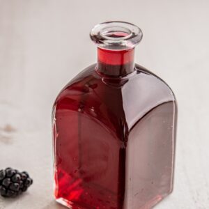 A clear glass bottle filled with a dark red liquid, possibly juice or syrup, sits on a light-colored surface next to a single blackberry.