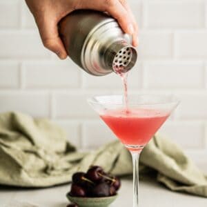 A person pours a pink cocktail from a shaker into a martini glass. A small bowl of cherries is in the background on a table with a green cloth. The scene has a light, airy atmosphere.