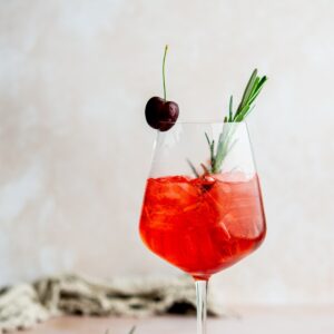 A stemmed glass filled with a cherry spritz garnished with ice, a sprig of rosemary, and a cherry on top. The background is soft and neutral, creating a refreshing and elegant presentation.