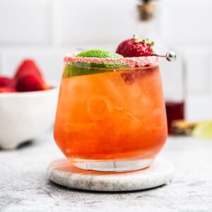 A refreshing strawberry margarita on the rocks in a glass with a sugared rim, garnished with a lime wedge and a strawberry on a skewer. The drink is orange-pink and served on a stone coaster. A bowl of strawberries and a bottle are blurred in the background.