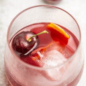 Close-up of a glass filled with a cherry old fashioned cocktail containing ice cubes, a fresh cherry with stem, and small slices of orange. The drink appears refreshing, with a lightly frosted exterior, suggesting its chilled.