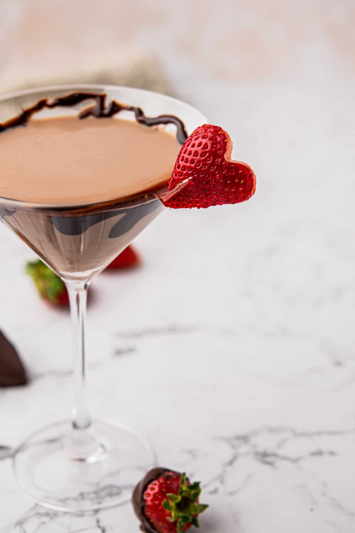 A chocolate martini in a glass with chocolate drizzle on the rim, garnished with a heart-shaped strawberry slice. Additional strawberries and chocolate pieces are scattered on a marble surface in the background.