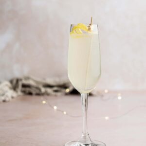 A tall glass brimming with a light, sparkling French 75 is garnished with a lemon twist on a skewer. The elegant cocktail rests on a pale surface, with soft, blurred lights in the background, creating an effortlessly refreshing scene.