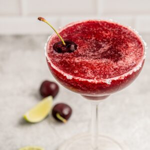 A vibrant frozen cherry margarita in a sugar-rimmed glass is garnished with two cherries, while more cherries and lime slices decorate the blurred background on a gray surface.