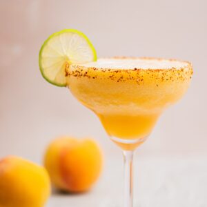 A frosty peach margarita with a chili-rimmed glass, garnished with a lime slice. Two peaches are blurred in the background against a soft beige backdrop.