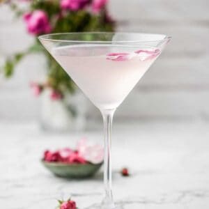 A martini glass filled with a pale pink rose cocktail, garnished with rose petals. The background is blurred, featuring pink flowers in a vase on a white surface.