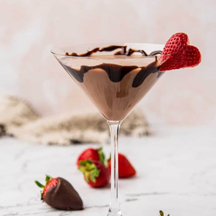 A martini glass filled with a creamy chocolate cocktail, drizzled with chocolate syrup. A halved strawberry garnishes the rim. Two strawberries, one chocolate-covered, rest on a marble surface nearby. Neutral background.