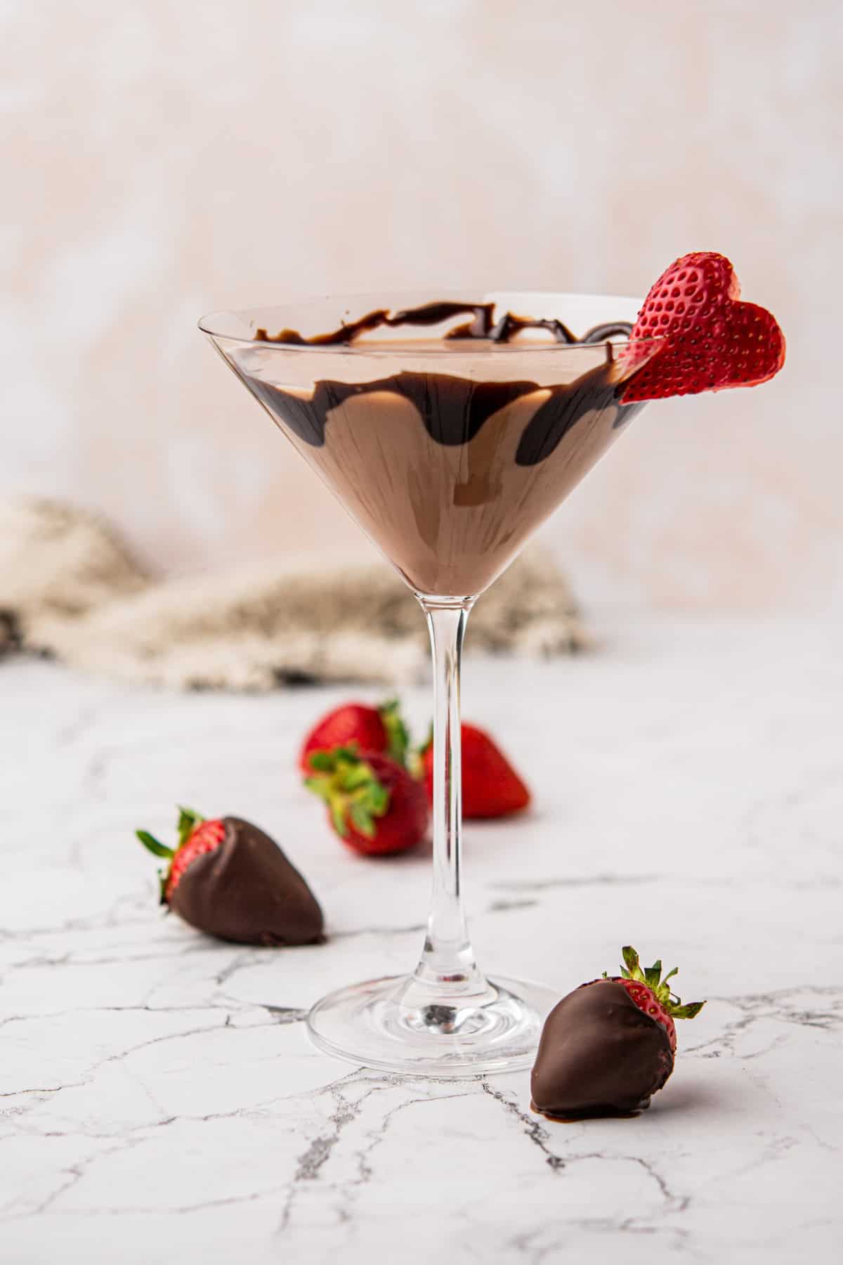 A martini glass filled with chocolate mousse, drizzled with chocolate syrup, garnished with a heart-shaped strawberry slice. Two chocolate-covered strawberries are placed on a marble surface nearby.
