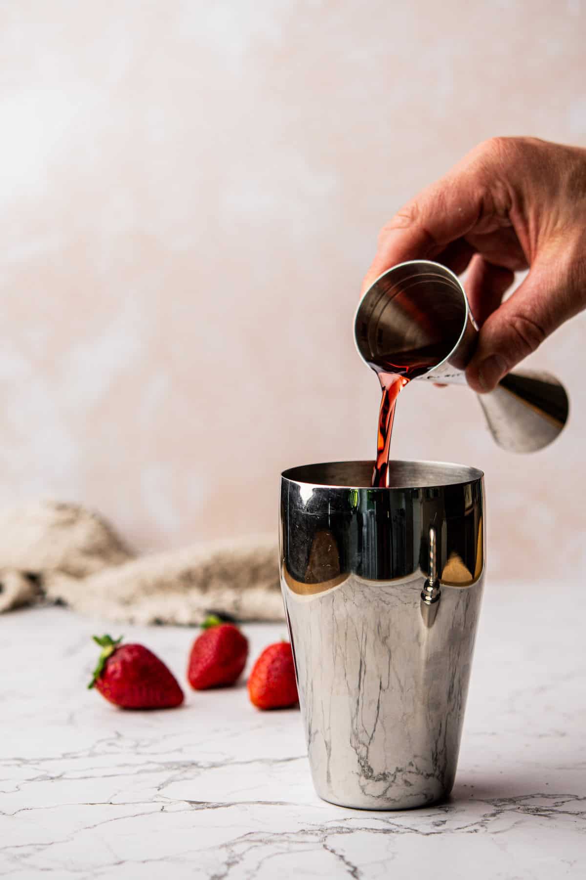 A hand pours liquid from a jigger into a stainless steel cocktail shaker on a marble surface. Three strawberries are nearby, and a beige cloth is in the background against a light pink, textured wall.