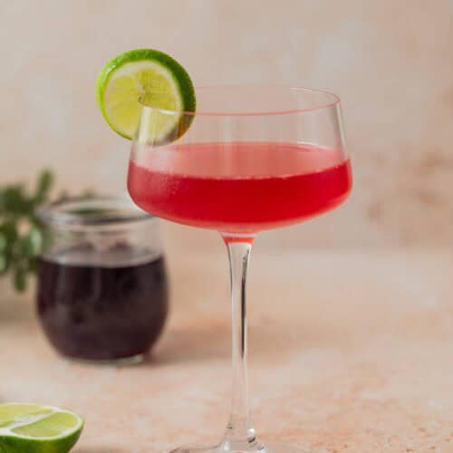A stemmed glass filled with a bright red cherry gimlet, garnished with a lime slice on the rim. In the background are a jar of dark liquid, a lime half, and a bowl of dark cherries.