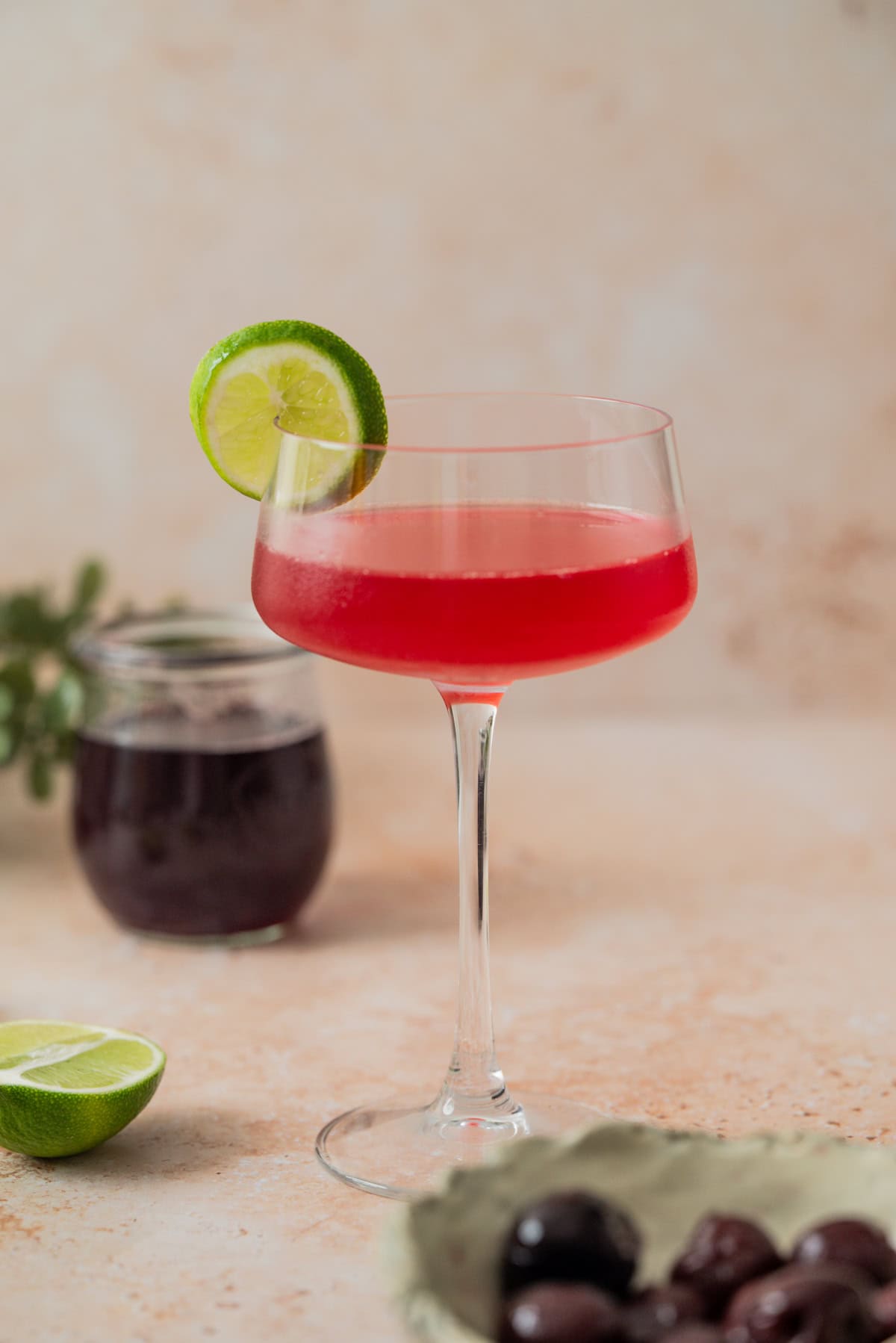 A stemmed glass filled with a bright red cherry gimlet, garnished with a lime slice on the rim. In the background are a jar of dark liquid, a lime half, and a bowl of dark cherries.