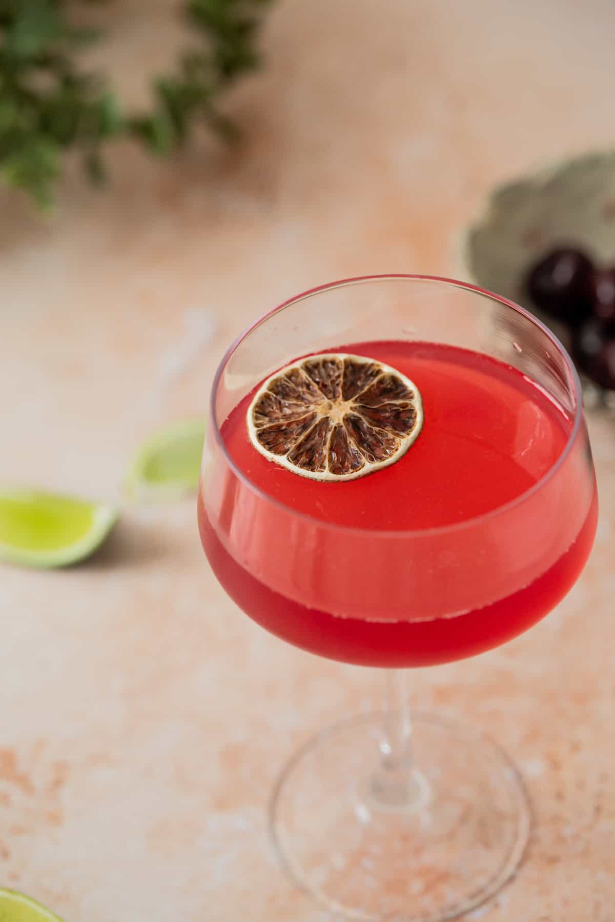 A cherry gimlet served in a stemmed glass, its vibrant red hue topped with a dried lime wheel, while lime wedges and cherries are softly blurred in the background.