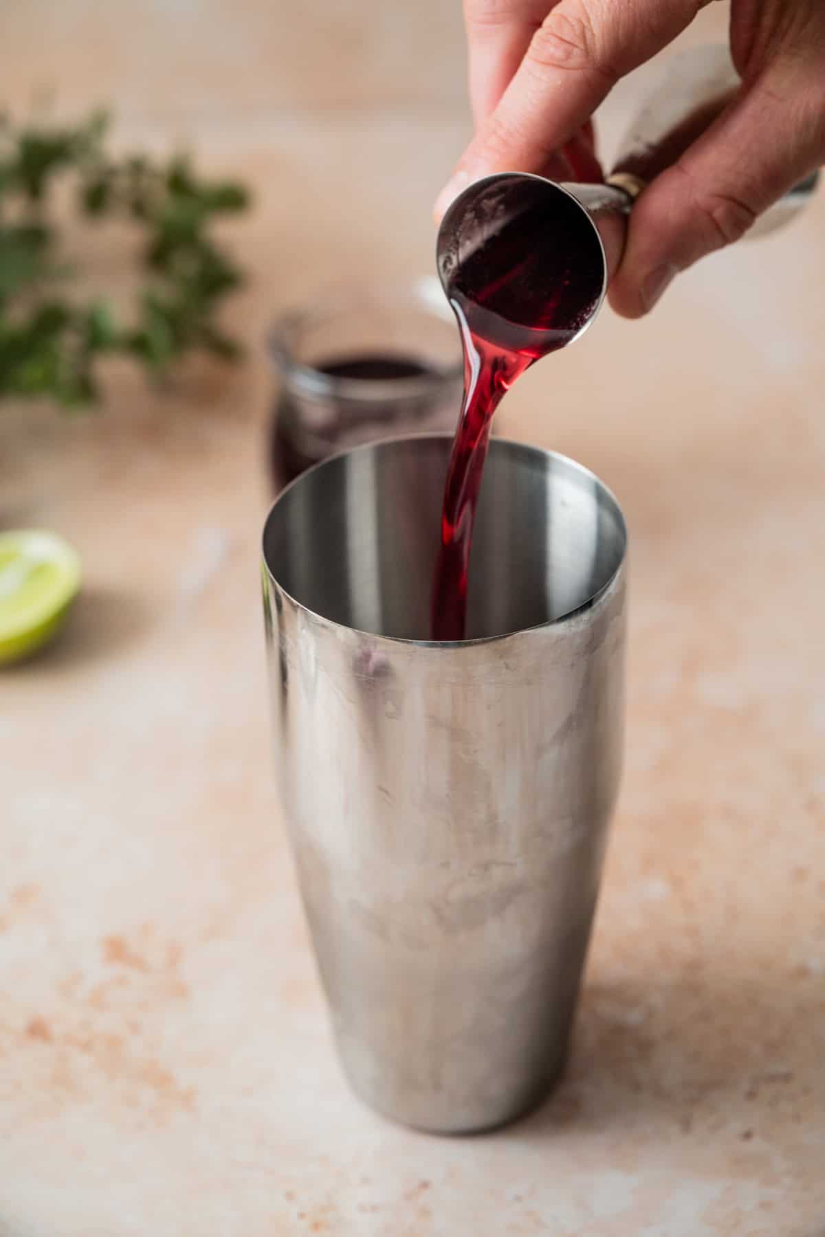 A hand pours deep red liquid for a cherry gimlet from a jigger into a metal cocktail shaker, with a jar of dark syrup, a lime half, and green herbs blurred in the background.