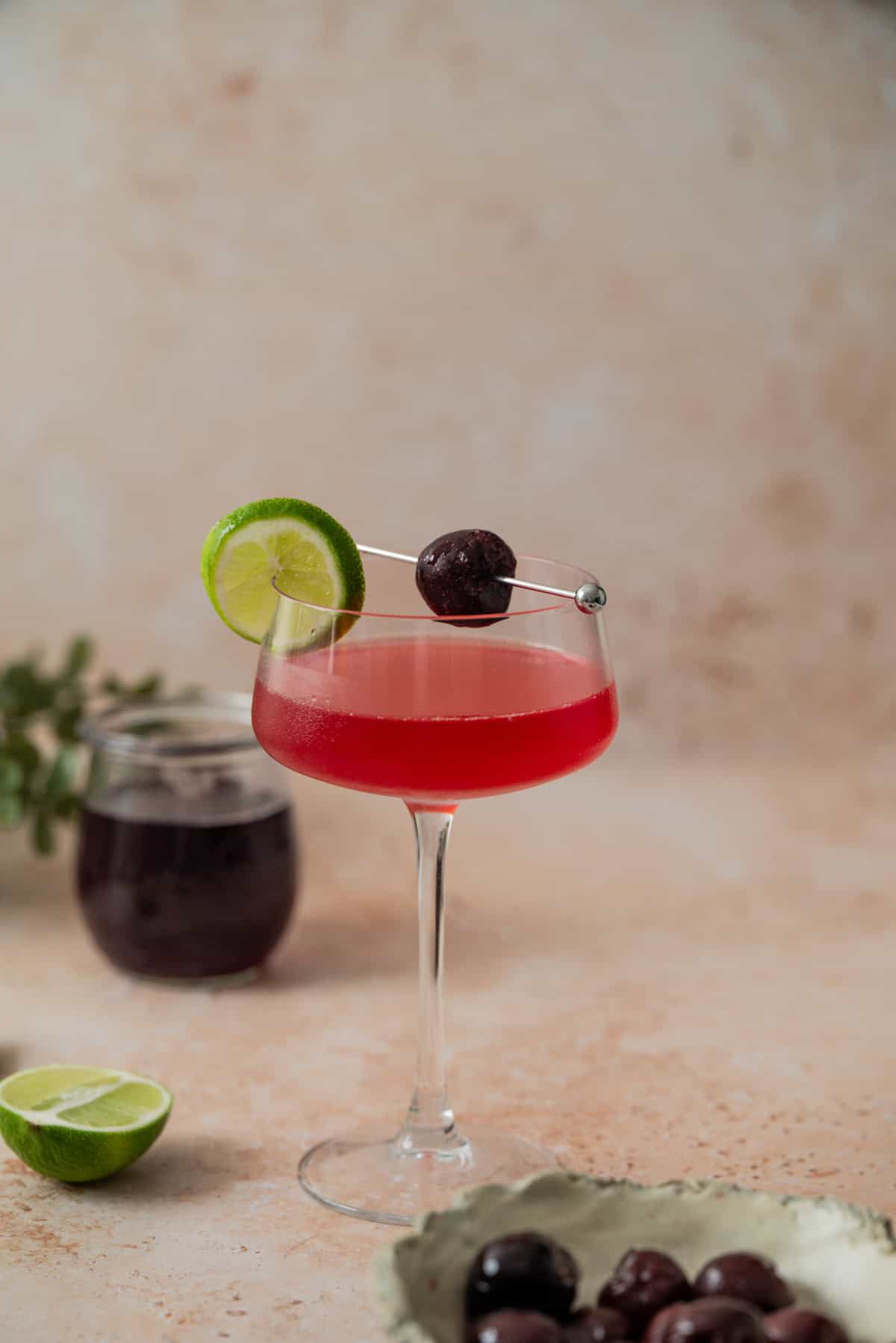 A coupe glass filled with a bright red cherry gimlet, garnished with a lime wheel and a dark cherry on a cocktail pick. A jar of dark liquid, lime halves, and a dish of cherries are in the background.