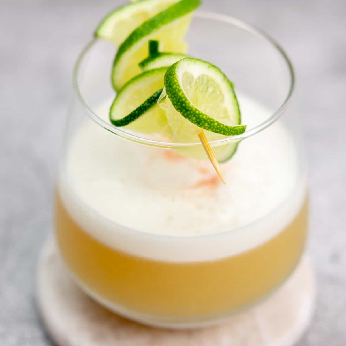 A frothy cocktail sits in a clear glass on a coaster. It is garnished with a skewer of spiral-cut lime slices. The drink has a creamy white foam on top and a light golden-brown liquid base. The background is softly blurred.