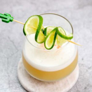 A tequila sour cocktail with a frothy top served in a round glass on a coaster, garnished with a skewer of spiral-cut lime slices. The background is a gray textured surface.