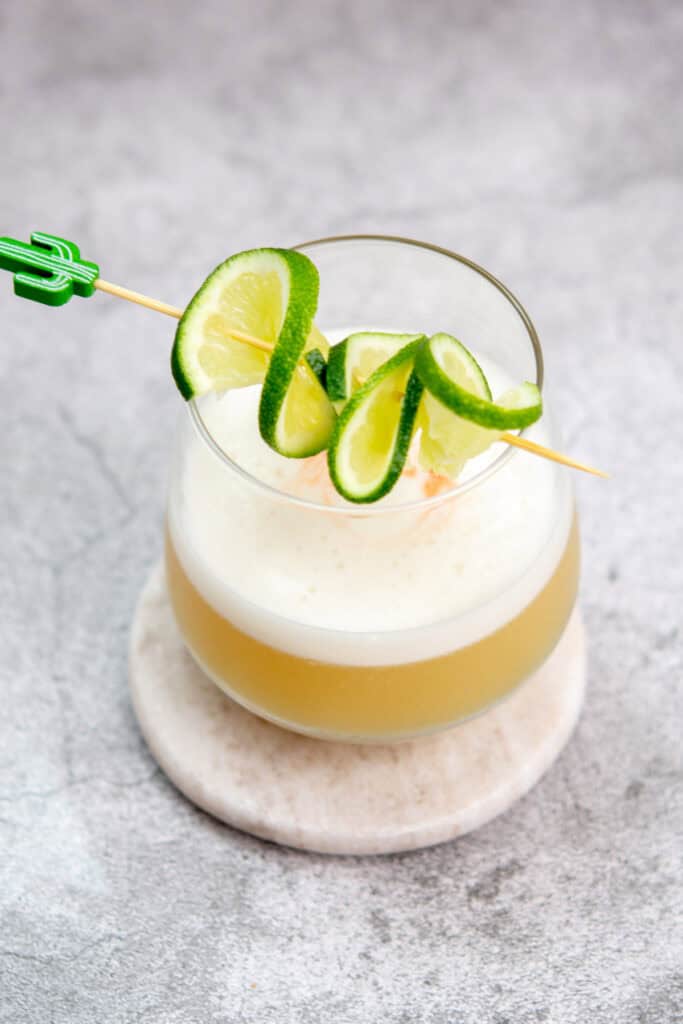 A cocktail in a round glass sits on a coaster. The drink has a frothy top and is garnished with slices of lime on a skewer. The background is a textured gray surface.