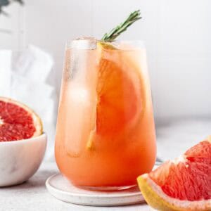 A glass of pink grapefruit cocktail with ice, a grapefruit slice, and a rosemary sprig garnish, sits on a coaster. Fresh grapefruit segments are on the table nearby.