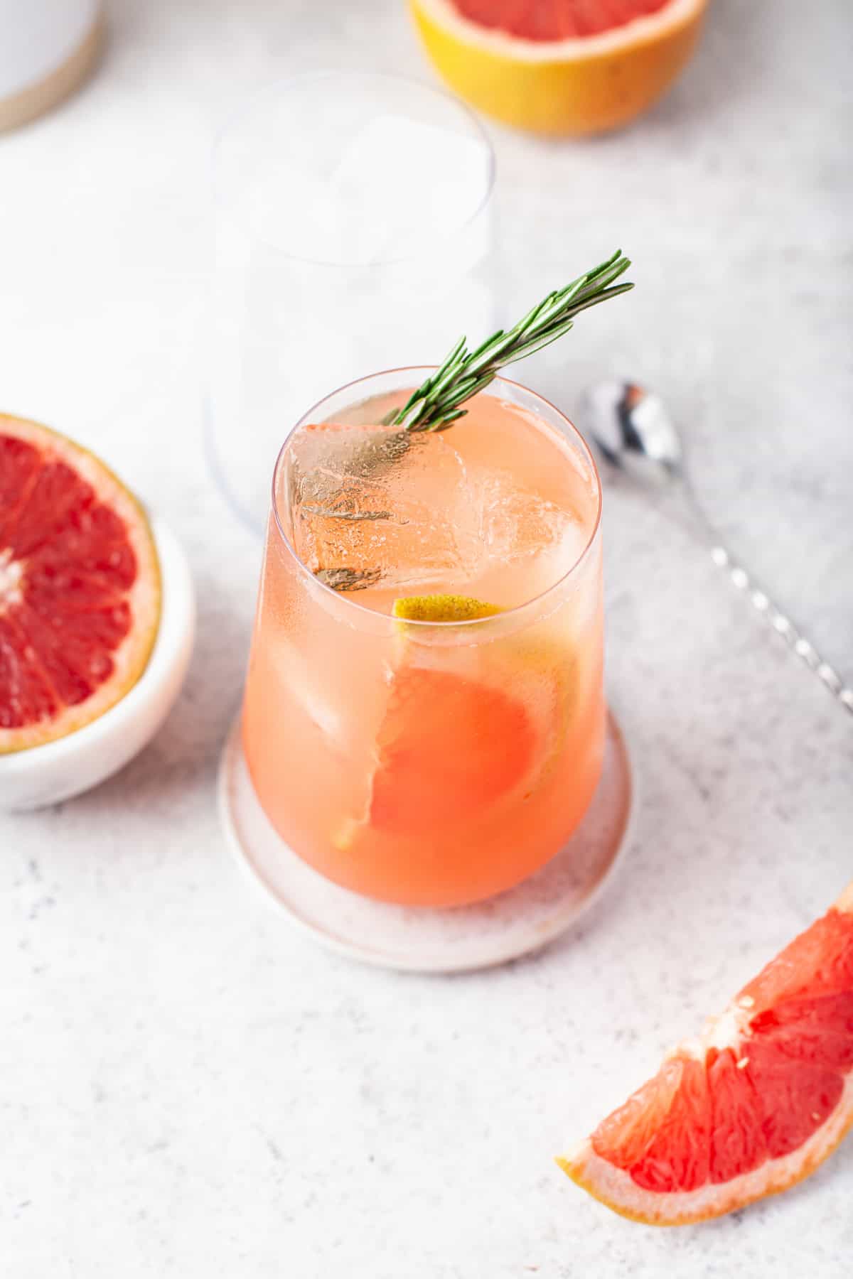 A glass of pink grapefruit cocktail with ice, a slice of grapefruit, and a sprig of rosemary, sits on a coaster. Fresh grapefruit halves and a wedge are nearby on a light surface.