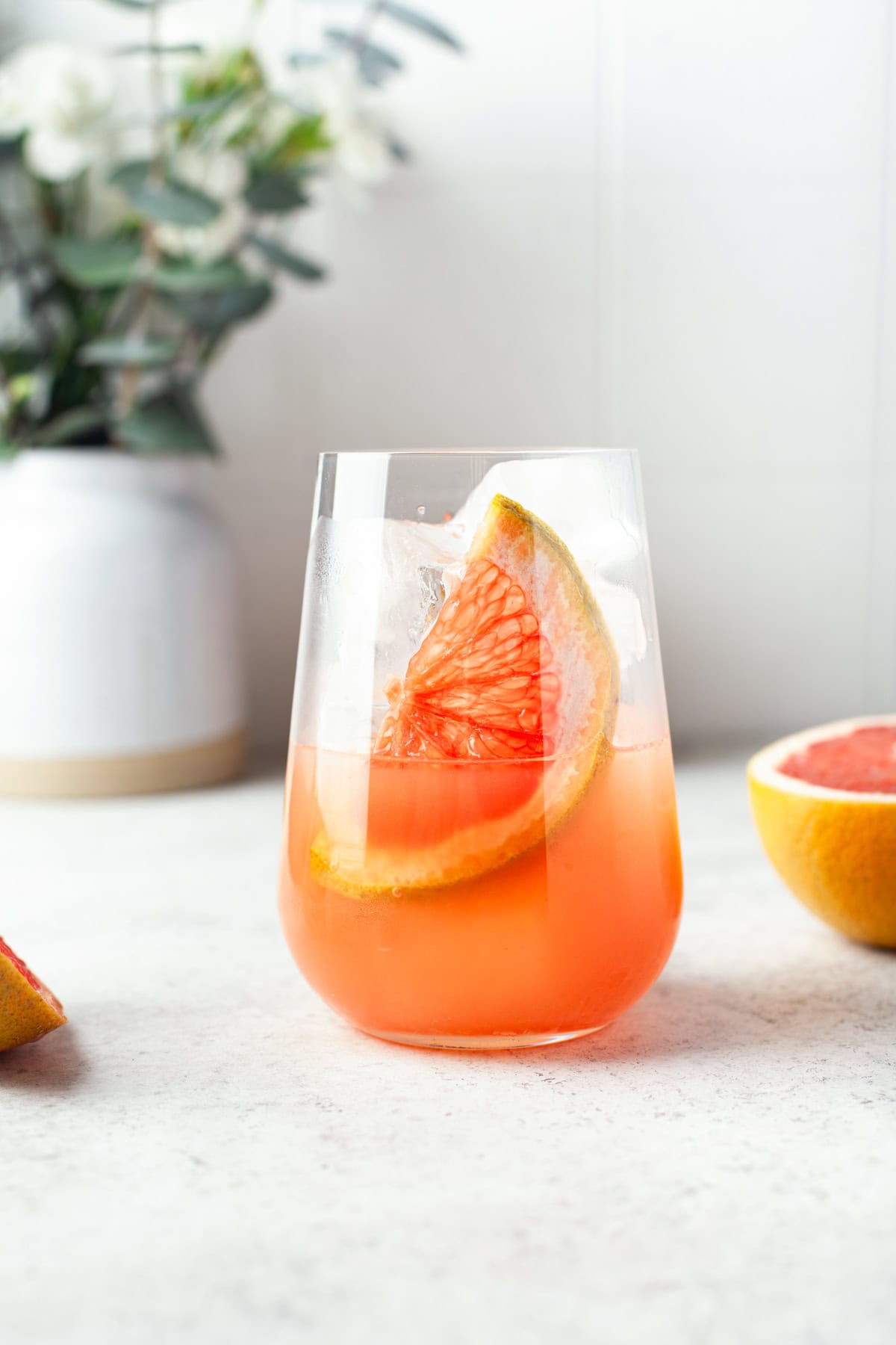 A glass of pink grapefruit cocktail with ice and a wedge of grapefruit, set on a light countertop. In the background, there is a halved grapefruit and a white vase with green foliage and small white flowers.