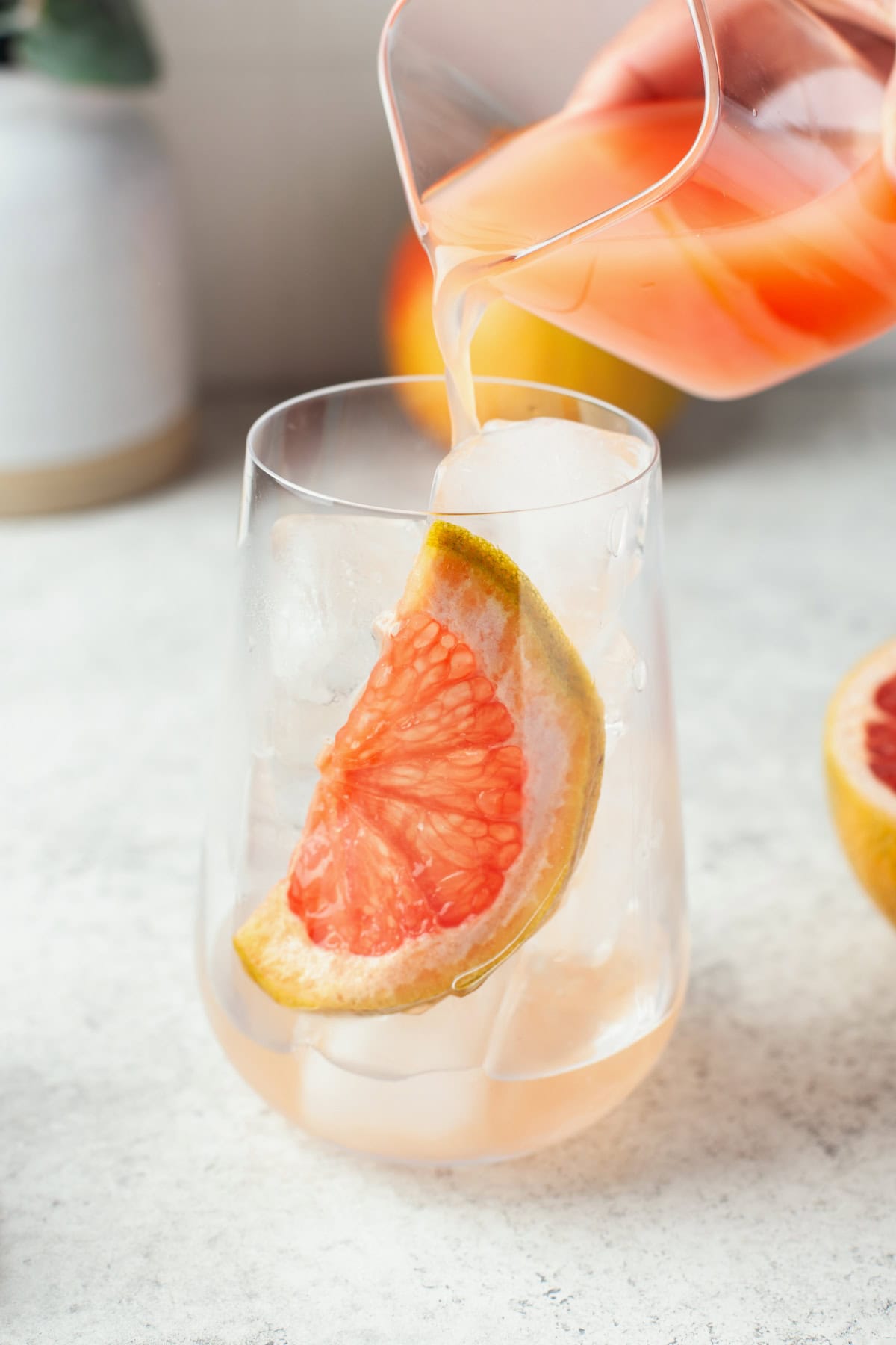 A hand pours pink grapefruit juice from a glass pitcher into a clear glass filled with ice and a large wedge of fresh grapefruit.