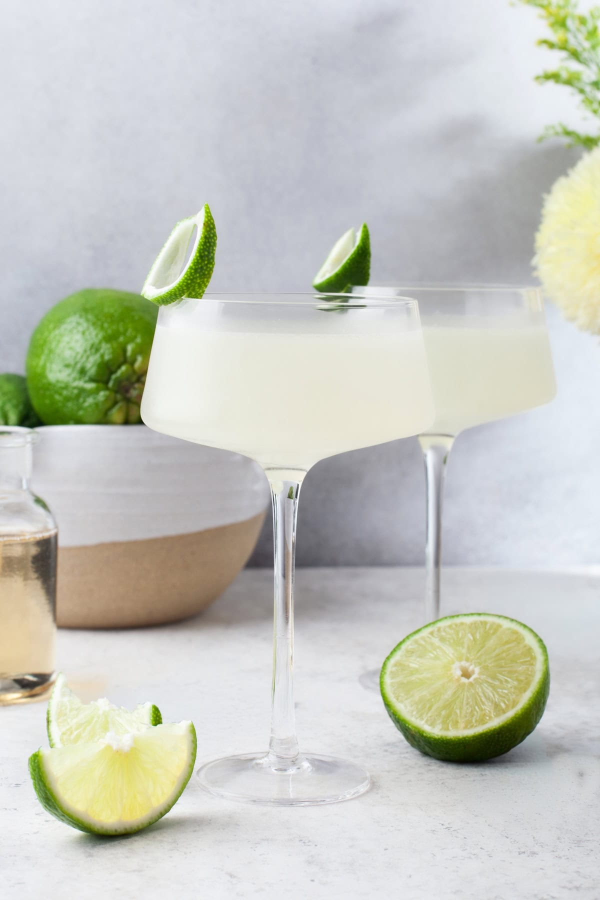Two coupe glasses filled with a pale green Vodka Gimlet, each garnished with a lime wedge. Fresh limes, a small bottle, and flowers are arranged on a light surface in the background.