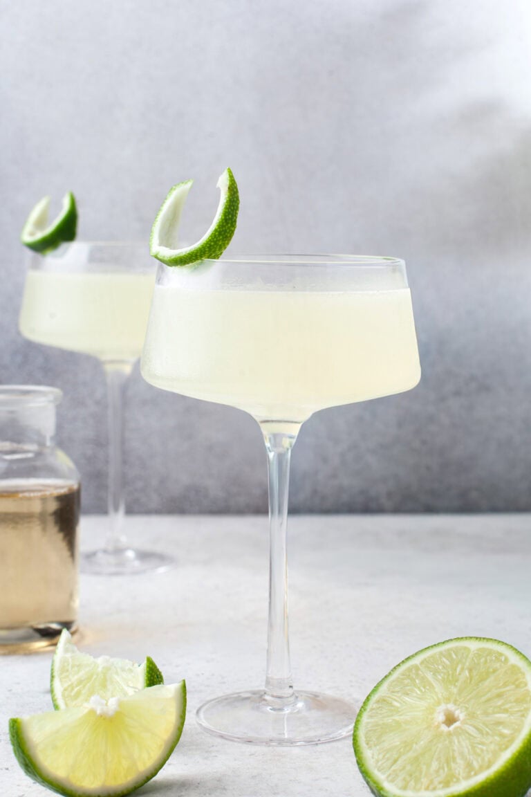 A glass of pale yellow cocktail garnished with a lime wedge sits on a light surface, with a second glass, a jar of liquid, and sliced limes in the background.