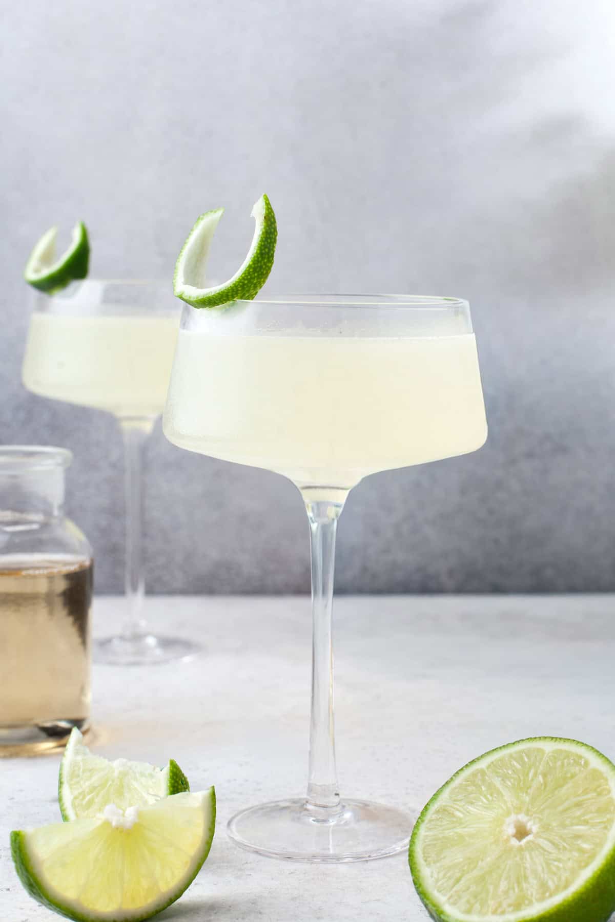 A glass of pale vodka gimlet garnished with a lime wedge sits on a light surface, with a second glass, a jar of liquid, and sliced limes in the background.
