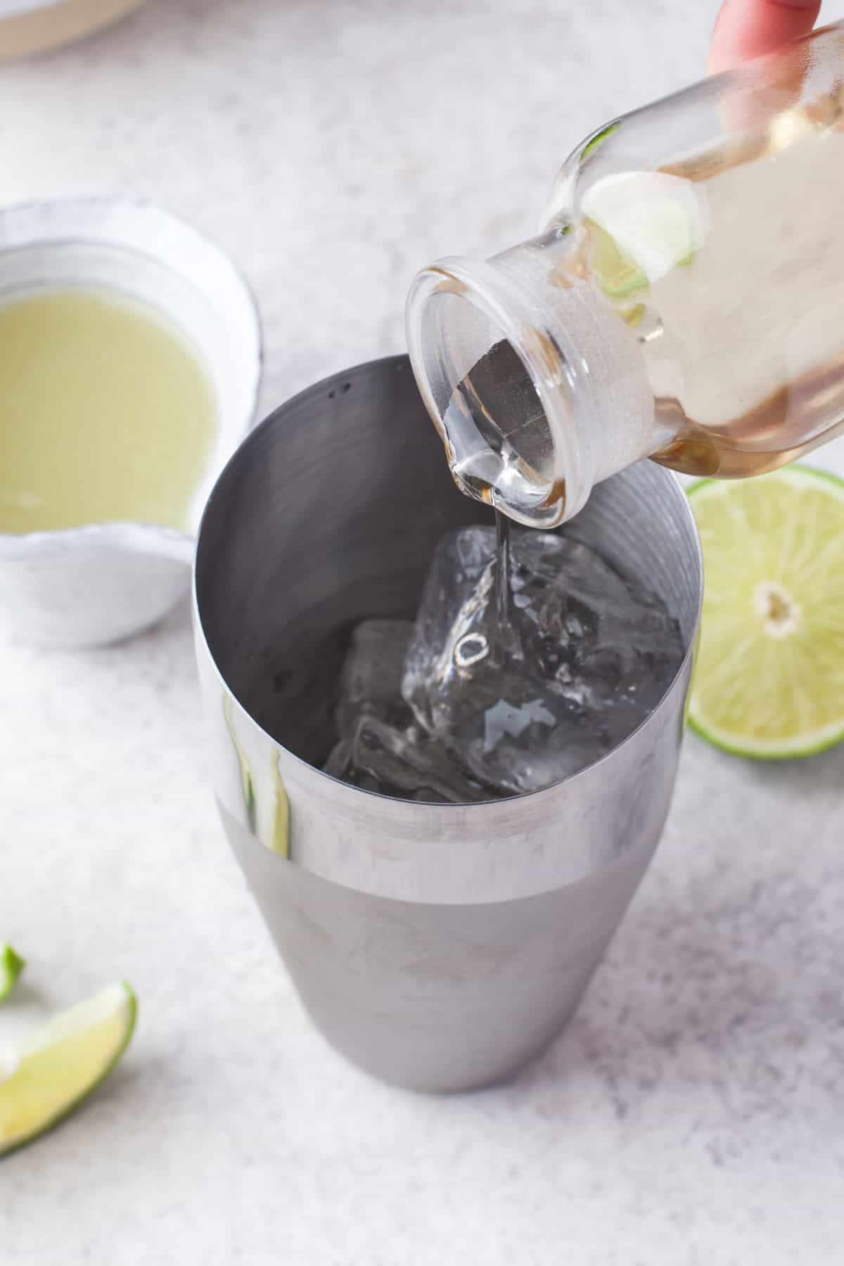 A hand pours clear liquid from a small bottle into a metal cocktail shaker filled with ice. Sliced limes and a bowl of lime juice are nearby on a light surface.
