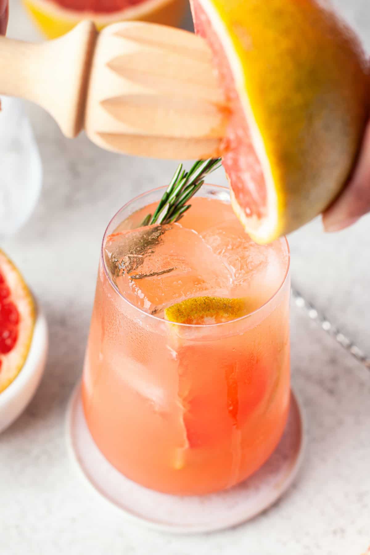 A close-up of a glass filled with pink grapefruit greyhound cocktail, ice, and a sprig of rosemary. A hand squeezes a fresh grapefruit over the drink using a wooden juicer.