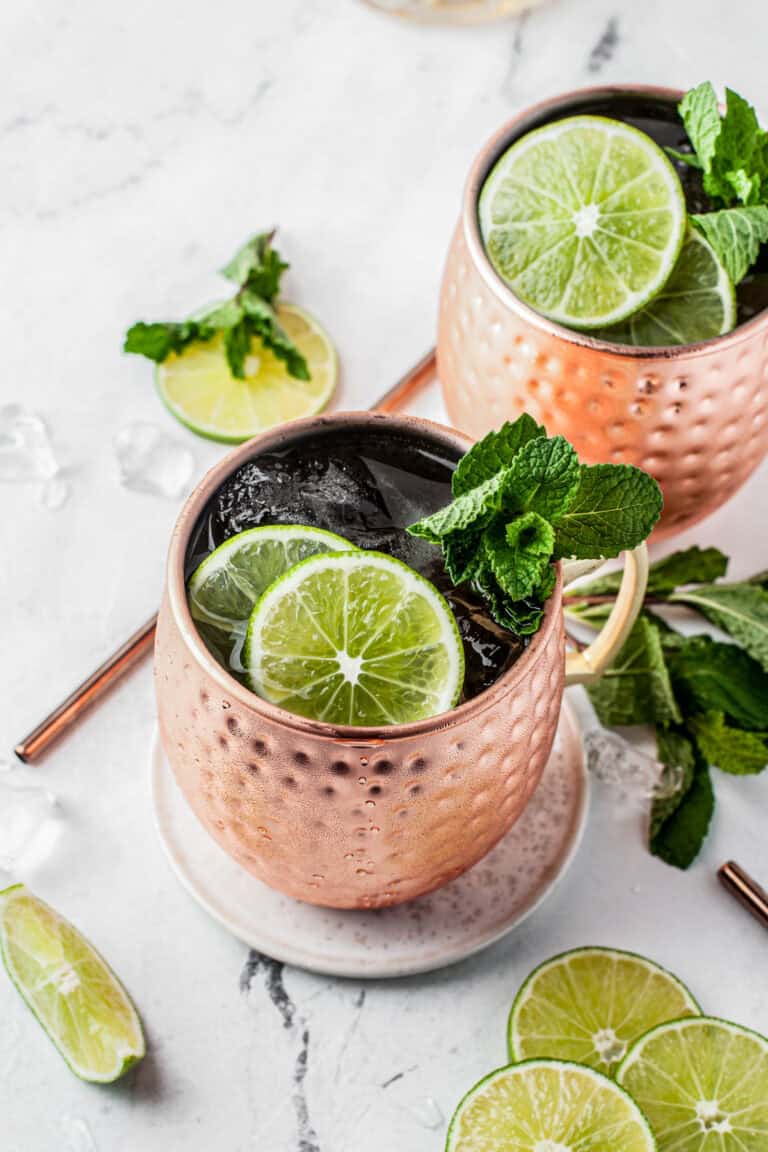 A copper mug filled with a cocktail, garnished with fresh lime slices and mint leaves, sits on a white coaster. Additional limes, mint, and another mug are in the background on a marble surface.
