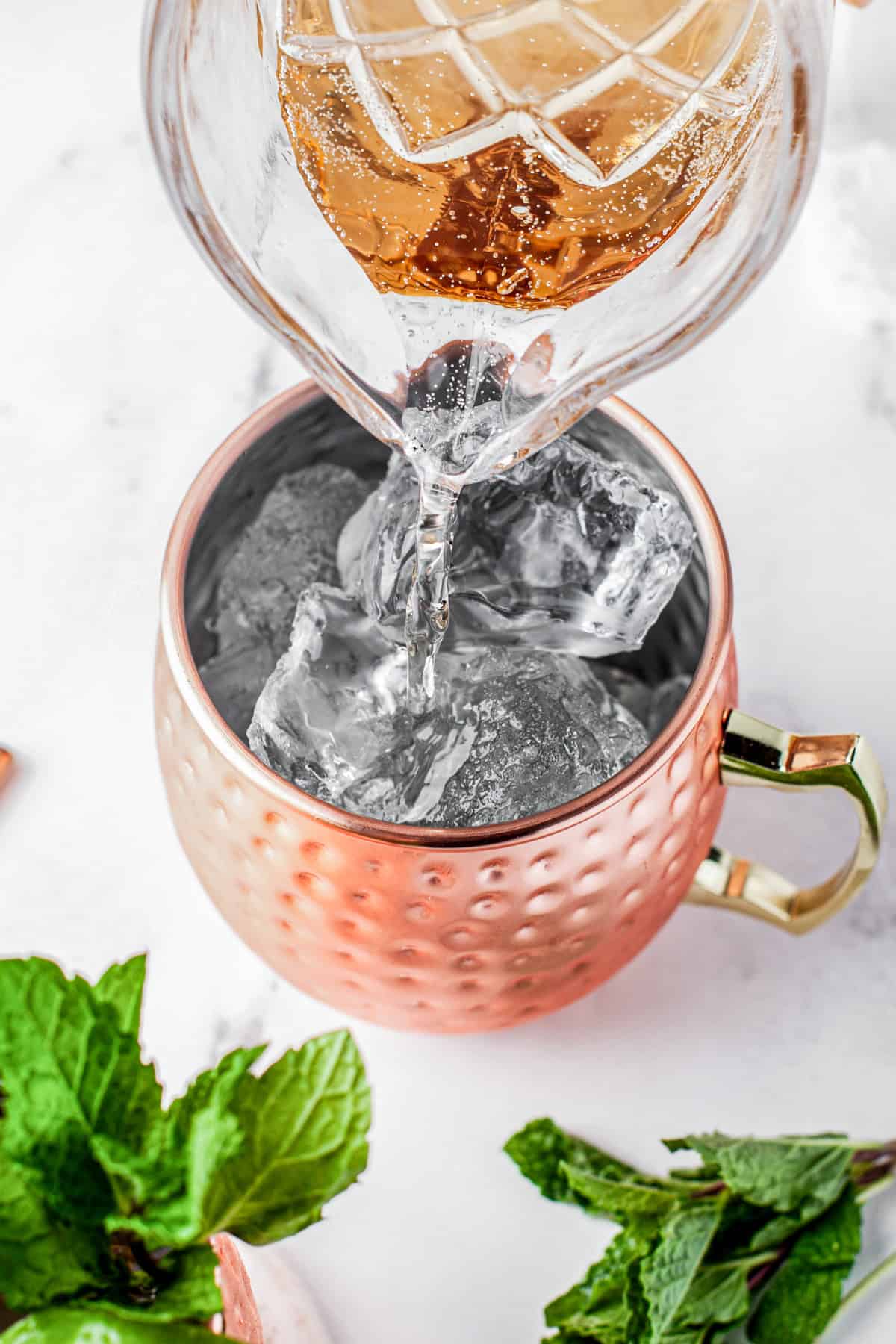 A glass pitcher pours a golden liquid over ice cubes in a copper mug with a handle. Fresh mint leaves are scattered nearby on a white surface.