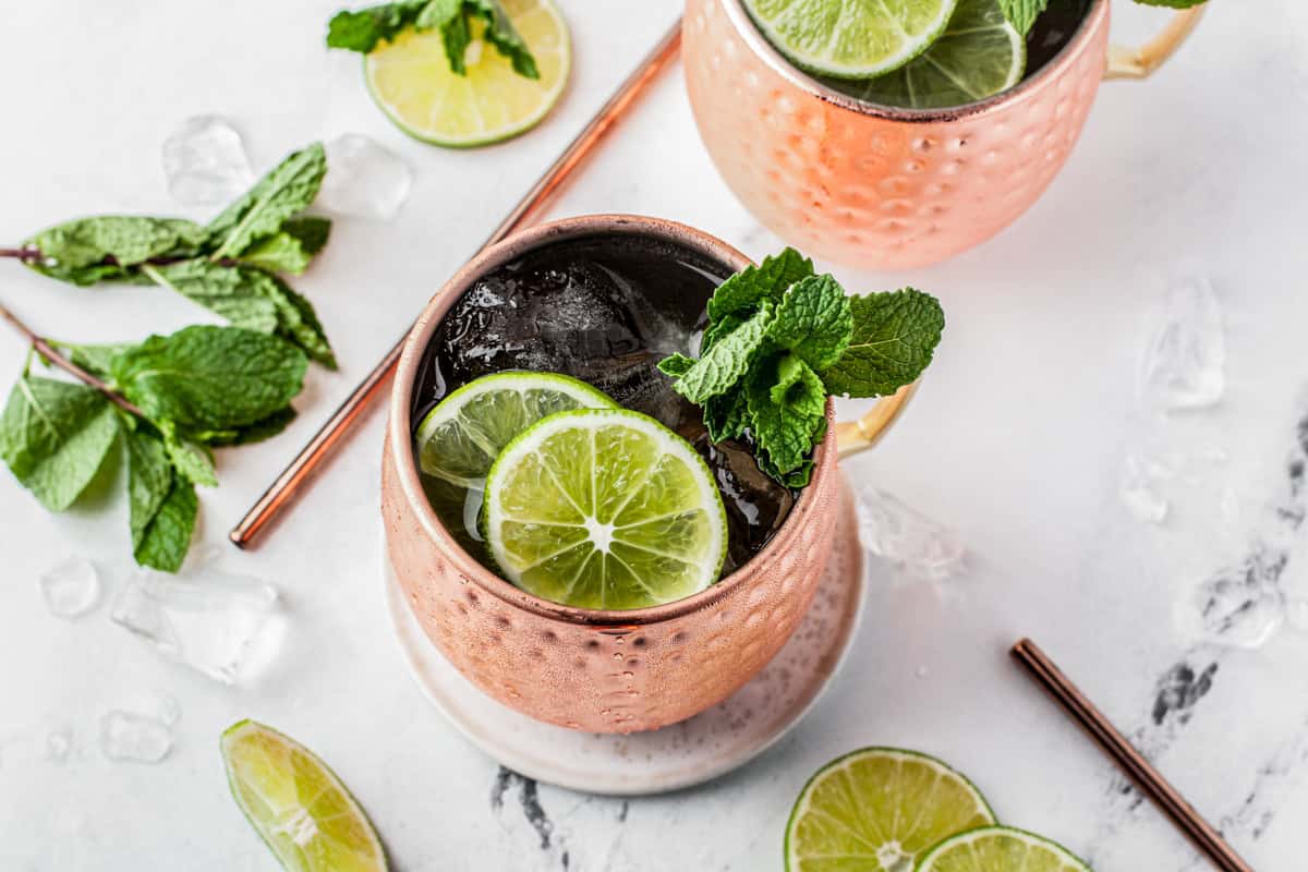 A copper mug filled with a cocktail, garnished with lime slices and fresh mint, sits on a white surface with ice cubes, mint sprigs, and lime wedges scattered around. Another similar drink is partially visible nearby.