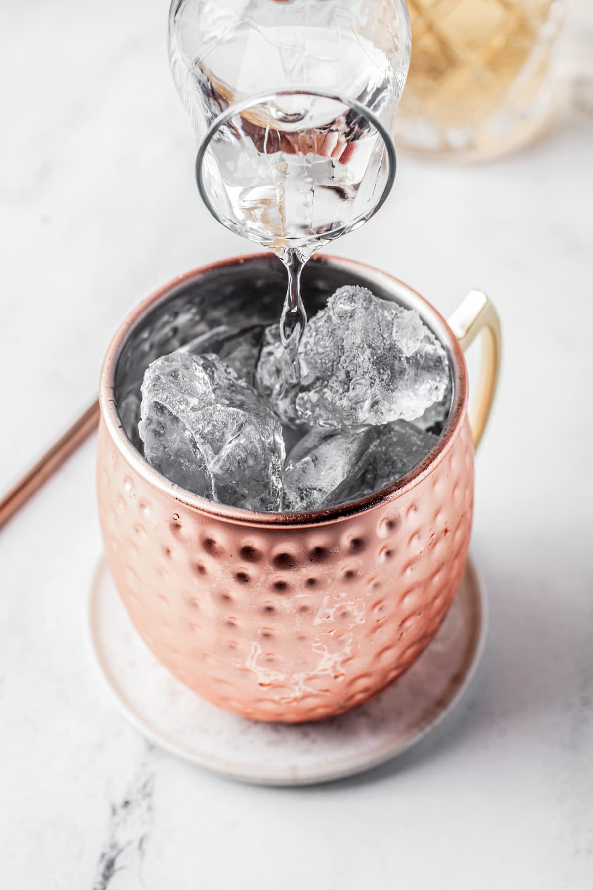 A clear liquid is being poured from a glass into a copper mug filled with ice cubes, sitting on a small white plate on a marble surface.