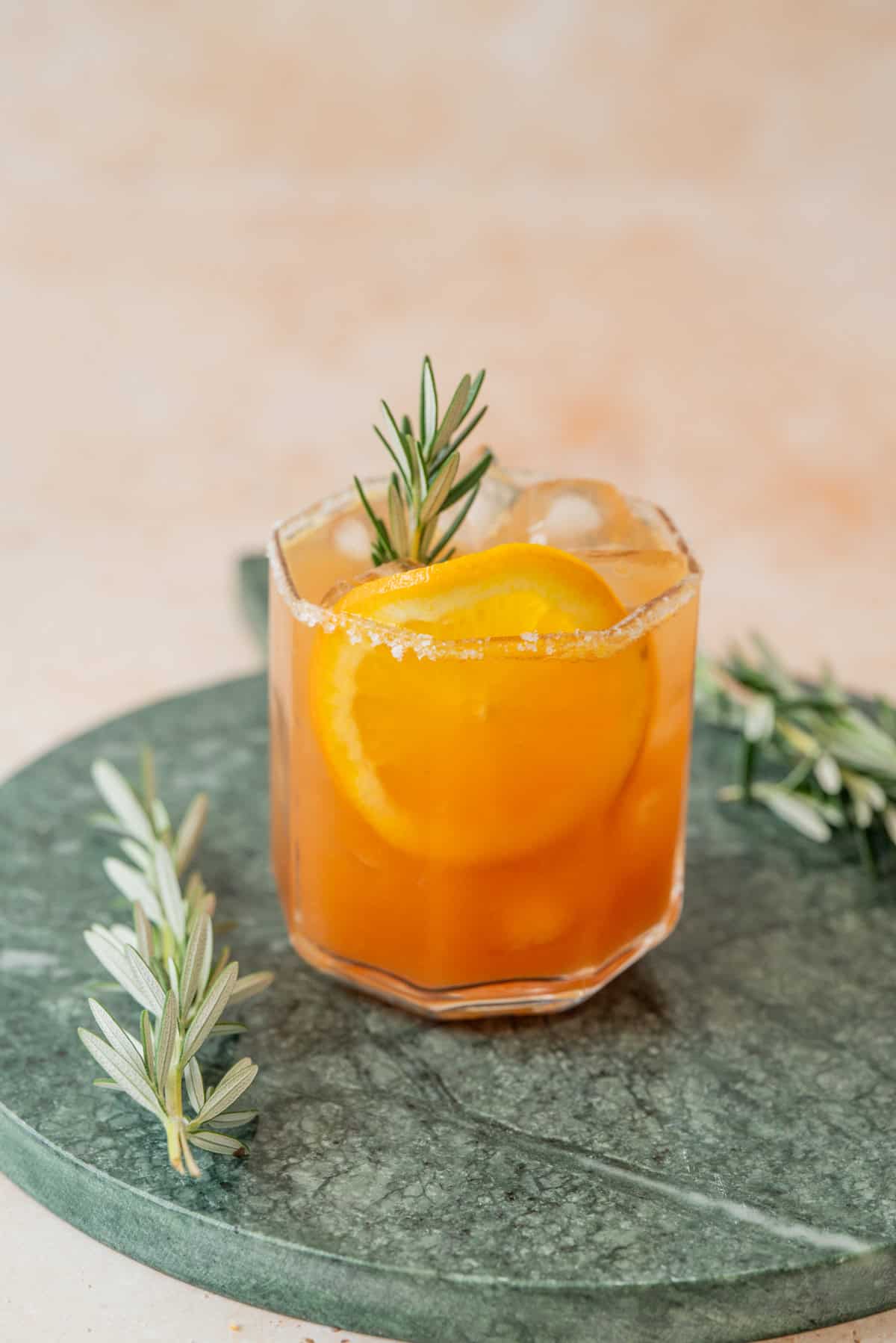 A glass of iced orange mocktail garnished with a sprig of rosemary and an orange slice, sits on a green marble surface with additional rosemary sprigs nearby.