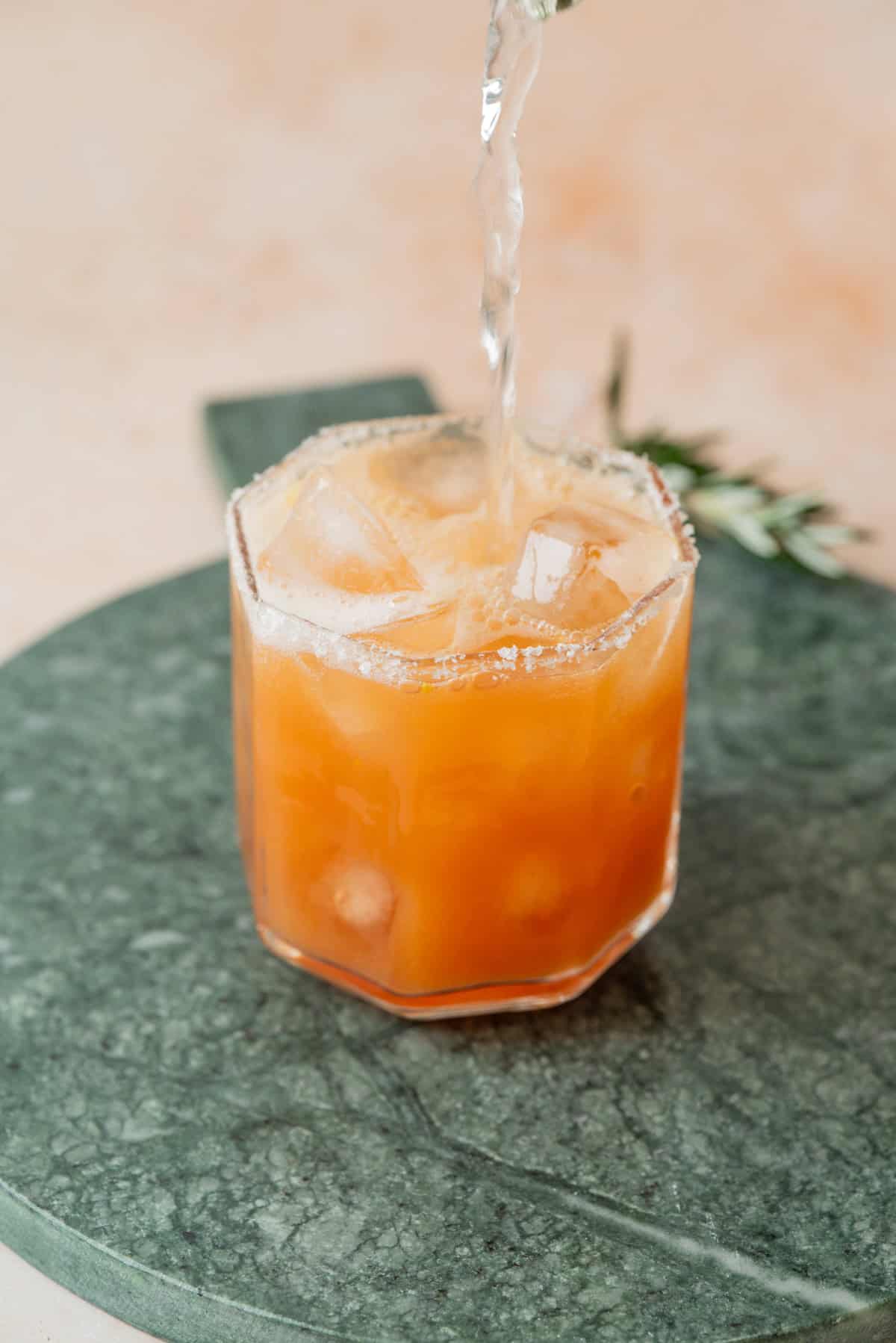 A glass of orange-colored iced Cranberry Orange & Rosemary Tonic, with a salted rim, is being filled with liquid. The glass sits on a green marble surface, with a sprig of rosemary in the background.